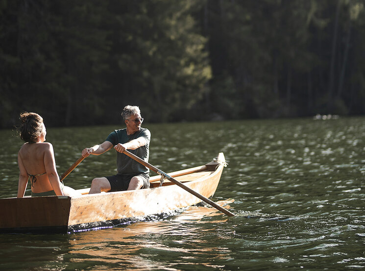 Zwei Menschen sitzen in einem hölzernen Ruderboot auf einem ruhigen See, umgeben von hohen Bäumen und üppigem Wald. Sonnenlicht spiegelt sich auf dem Wasser, während eine der beiden Personen das Boot rudert.