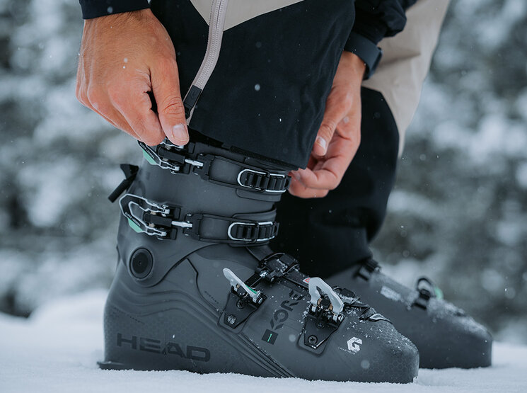 Eine Person steht im Schnee und stellt die Schnallen an einem Paar schwarzer HEAD-Skischuhe ein, während im unscharfen Hintergrund schneebedeckte Bäume zu sehen sind.