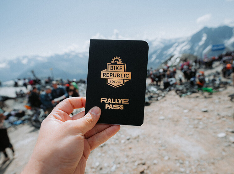 Eine Hand hält im Freien ein „Bike Republic Rallye Pass“-Heft in der Hand; im Hintergrund sind felsiger Boden und eine Menschenmenge zu sehen, während in der Ferne schneebedeckte Berge und ein strahlend blauer Himmel zu erkennen sind.
