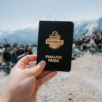 Eine Hand hält im Freien ein „Bike Republic Rallye Pass“-Heft in der Hand; im Hintergrund sind felsiger Boden und eine Menschenmenge zu sehen, während in der Ferne schneebedeckte Berge und ein strahlend blauer Himmel zu erkennen sind.