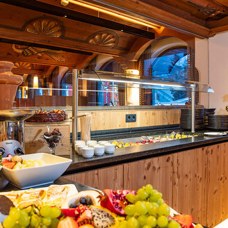 Ein Buffet aus Holz mit einem Schokoladenbrunnen, verschiedenen Obstsorten, Kuchen und Schalen in einem gemütlichen, warm beleuchteten Raum mit großen Fenstern und dekorativen Blumen.