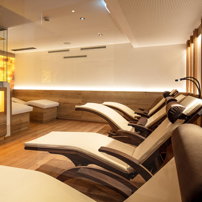 Moderner Wellness-Ruheraum mit mehreren gepolsterten Liegestühlen, sanfter Beleuchtung, Holzböden und einer warm beleuchteten Wand. Links ist eine verglaste Wand zu sehen, die eine ruhige, einladende Atmosphäre schafft.