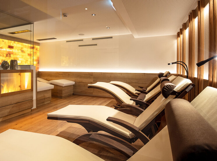Moderner Wellness-Ruheraum mit mehreren gepolsterten Liegestühlen, sanfter Beleuchtung, Holzböden und einer warm beleuchteten Wand. Links ist eine verglaste Wand zu sehen, die eine ruhige, einladende Atmosphäre schafft.