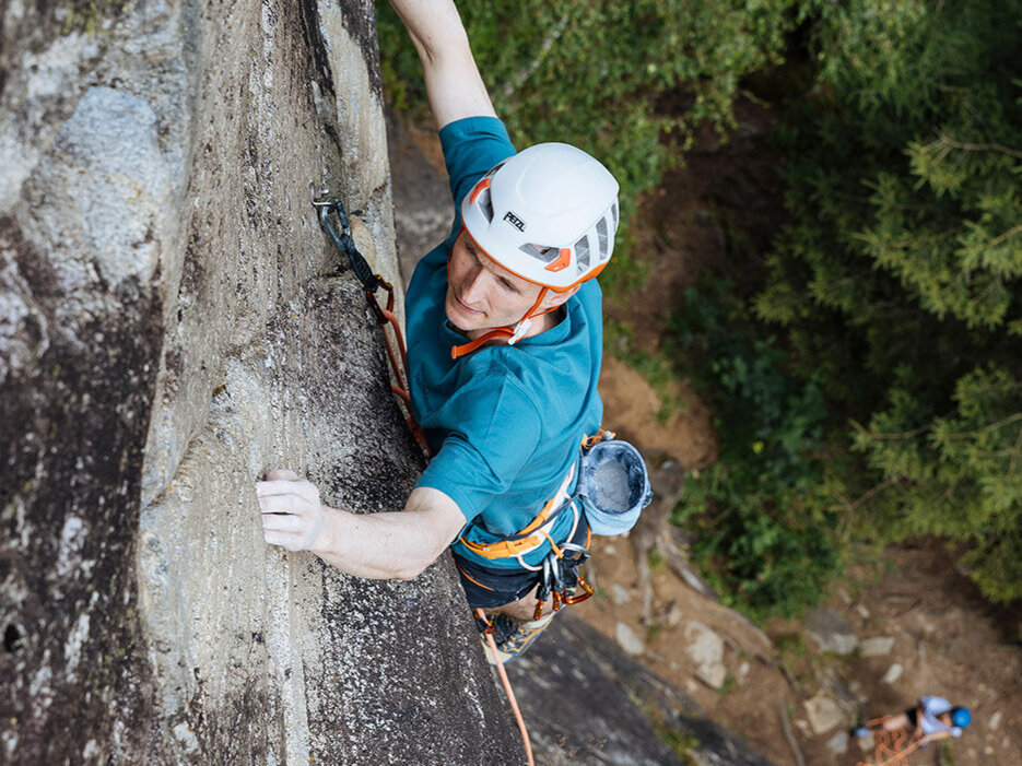 Eine Person, die einen Helm und einen Klettergurt trägt, klettert im Freien an einer steilen Felswand empor und greift dabei mit beiden Händen nach Tritten; unter ihr sind Bäume und ein weiterer Kletterer zu sehen.