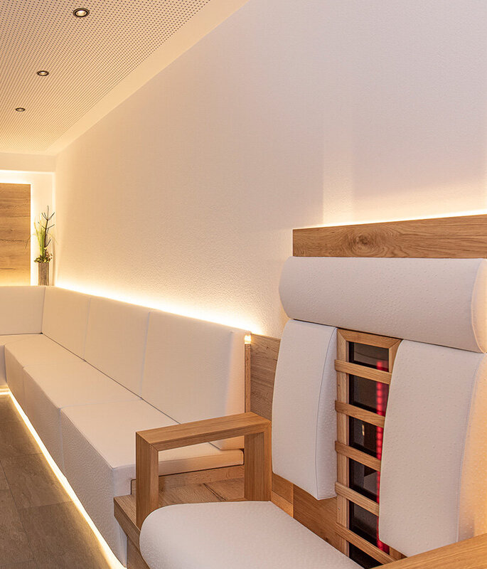 Eine moderne Infrarotlounge mit sanften, weißen Sitzbänken entlang der Wände, zwei runden Holztischen, einem Holzstuhl und minimalistischer Einrichtung. Warme LED-Beleuchtung unterstreicht die klaren Linien, und zwei kleine Topfpflanzen schmücken eine Holzverkleidung.