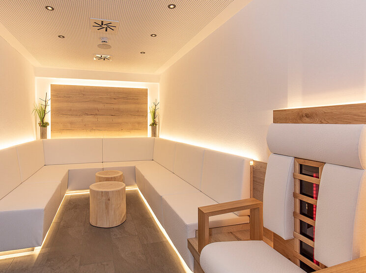 Eine moderne Infrarotlounge mit sanften, weißen Sitzbänken entlang der Wände, zwei runden Holztischen, einem Holzstuhl und minimalistischer Einrichtung. Warme LED-Beleuchtung unterstreicht die klaren Linien, und zwei kleine Topfpflanzen schmücken eine Holzverkleidung.