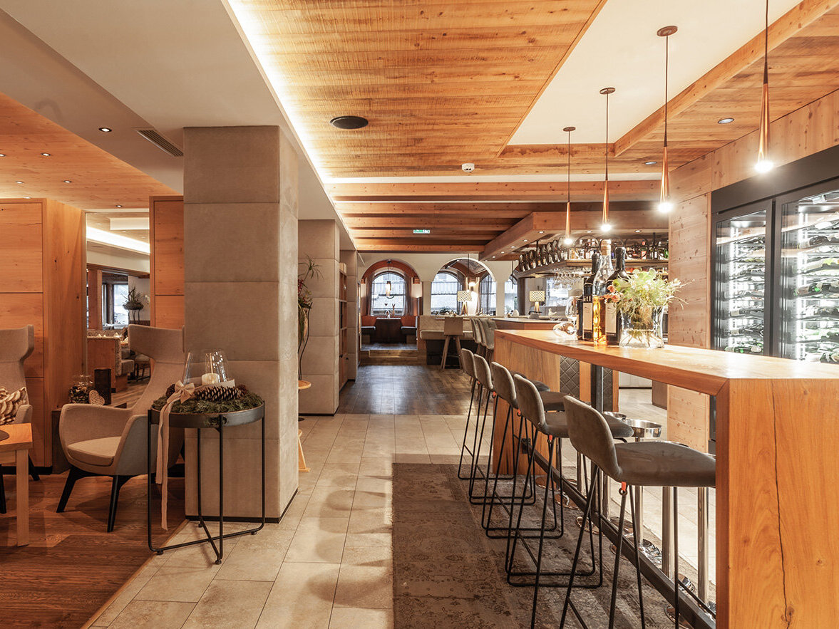 Modernes Restaurantinterieur mit Holzwänden und -decke, hohen Barhockern an der Theke, Loungesesseln, sanfter Beleuchtung und in einer Glasvitrine ausgestellten Weinflaschen. Die gesamte Atmosphäre ist warm und einladend.
