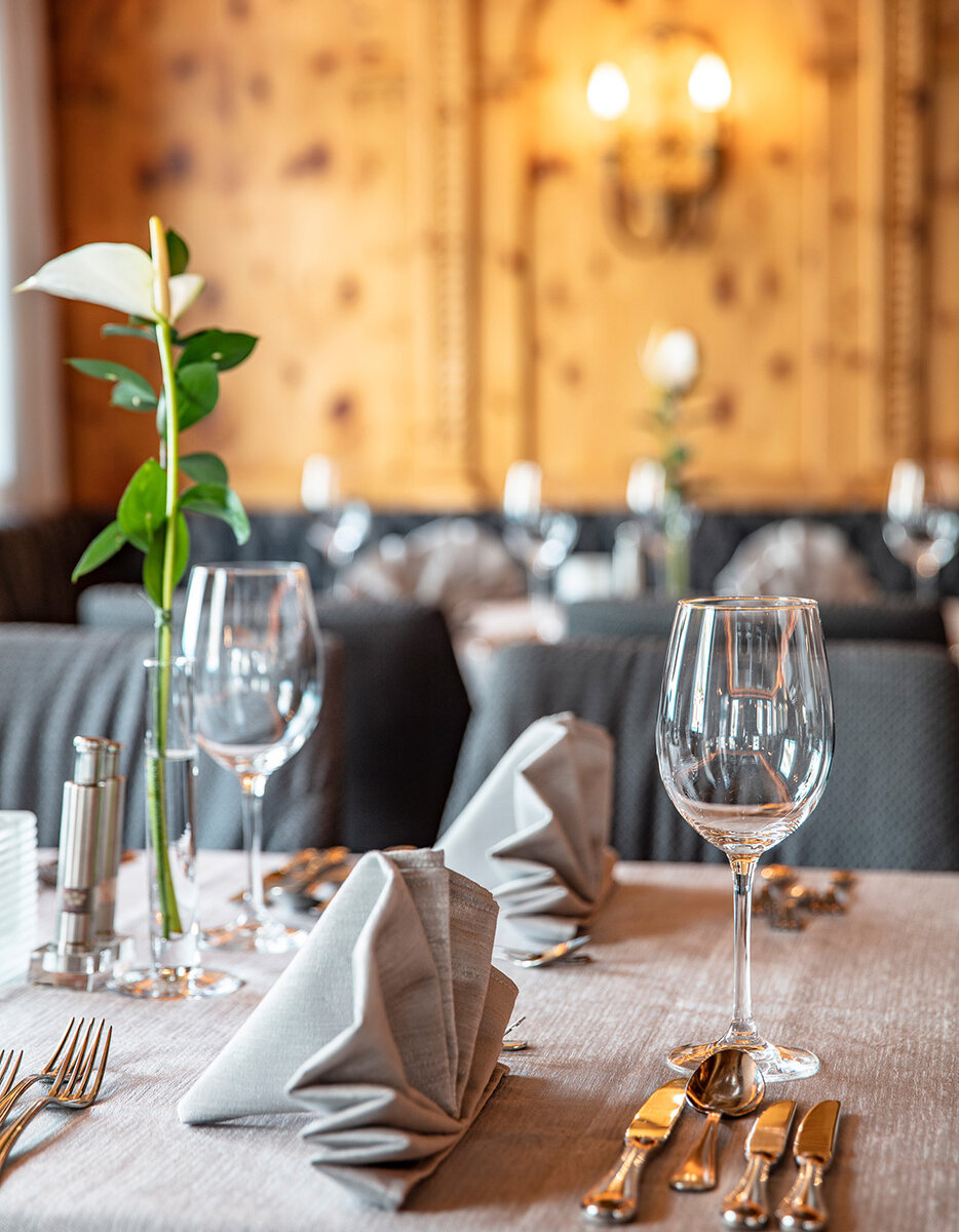 Ein elegantes Restaurant-Tischgedeck mit zwei Weingläsern, gefalteten grauen Servietten, Besteck und einer einzelnen weißen Blume in einer Vase. Der Hintergrund ist sanft beleuchtet, mit warmen Holzvertäfelungen und unscharf wirkenden Dekorelementen.