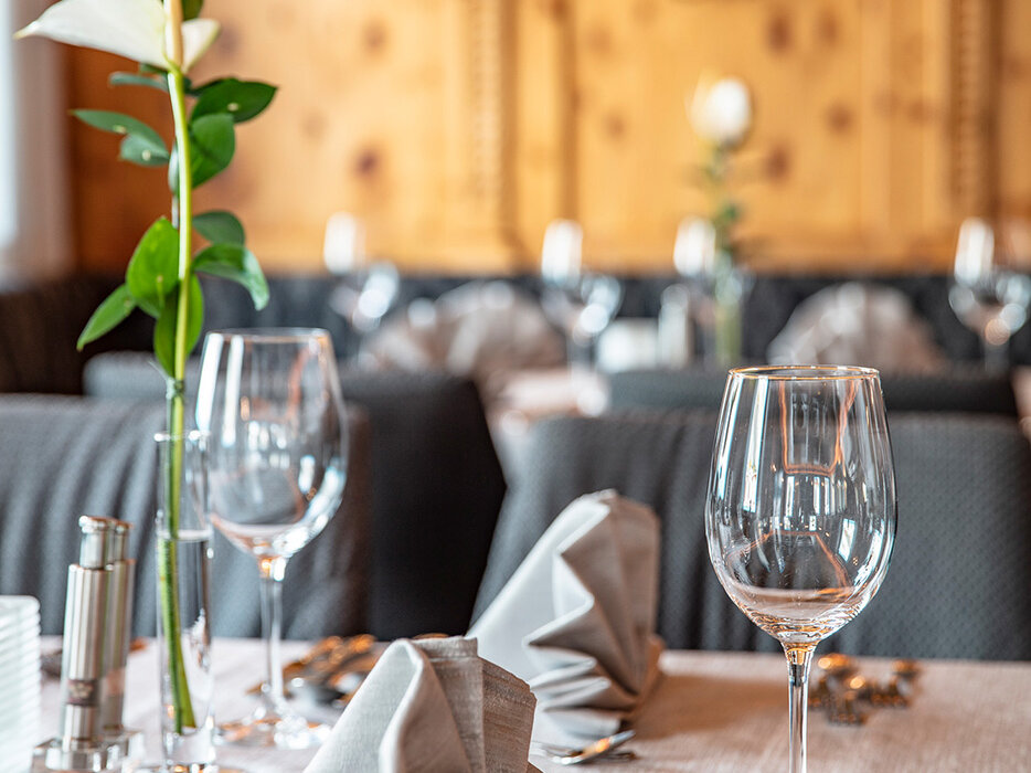 Ein elegantes Restaurant-Tischgedeck mit zwei Weingläsern, gefalteten grauen Servietten, Besteck und einer einzelnen weißen Blume in einer Vase. Der Hintergrund ist sanft beleuchtet, mit warmen Holzvertäfelungen und unscharf wirkenden Dekorelementen.