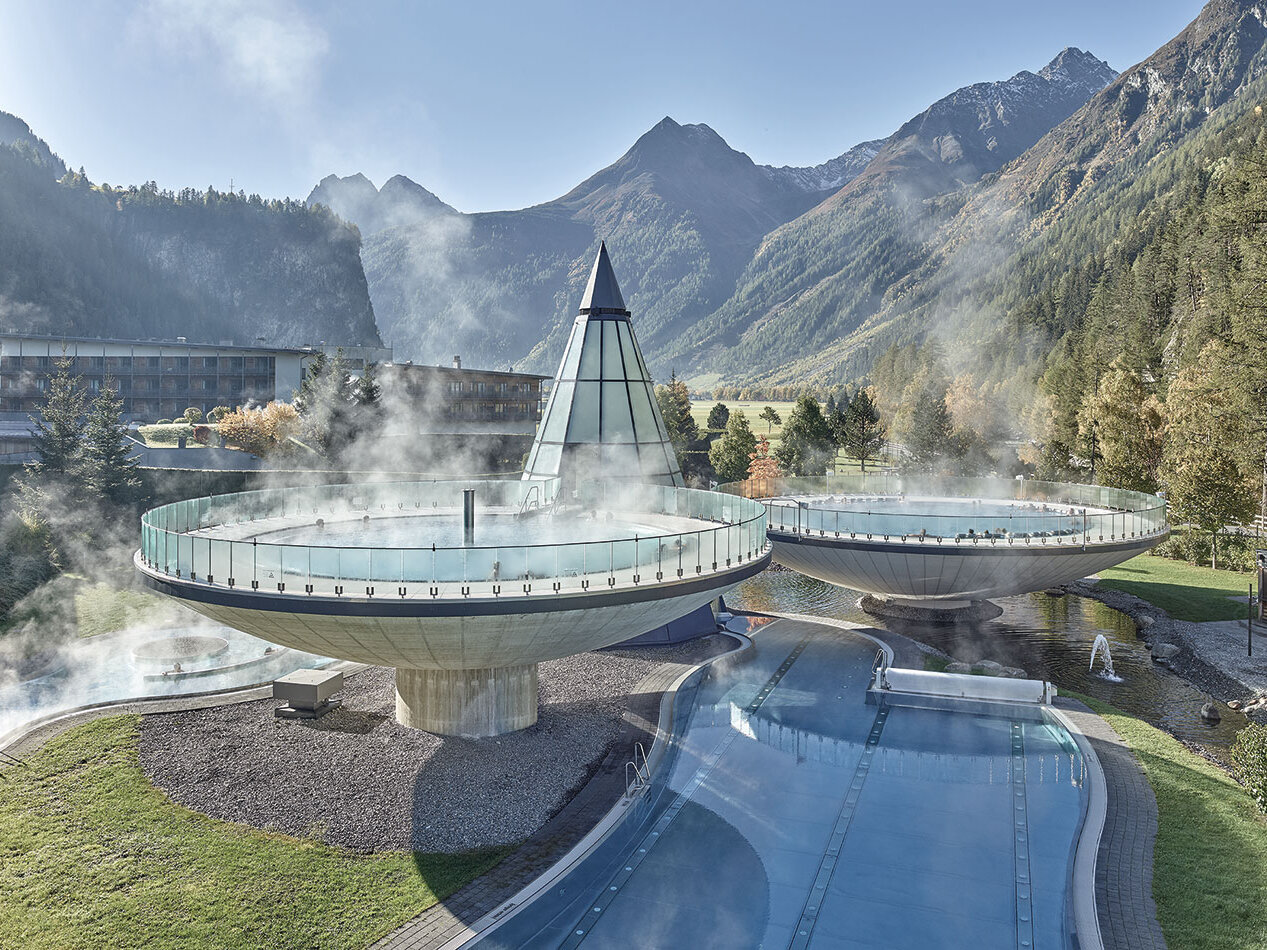 Thermalbäder im Freien mit aufsteigendem Dampf, Glasgeländern und einer spitz zulaufenden Glaskonstruktion, vor dem Hintergrund bewaldeter Berge und eines Tals unter klarem Himmel.