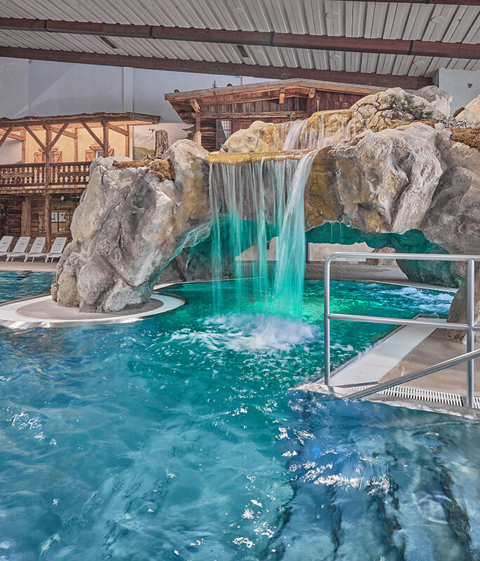 Hallenbad, Freizeit Arena, mit einer Felslandschaft, die einen Wasserfall und einen Tunnel unter den Felsen umfasst, sowie Liegestühlen, die in einem thematisch gestalteten, rustikalen Ambiente am Beckenrand aufgereiht sind.