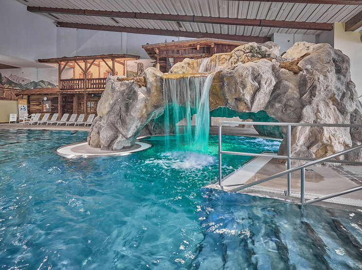 Hallenbad, Freizeit Arena, mit einer Felslandschaft, die einen Wasserfall und einen Tunnel unter den Felsen umfasst, sowie Liegestühlen, die in einem thematisch gestalteten, rustikalen Ambiente am Beckenrand aufgereiht sind.