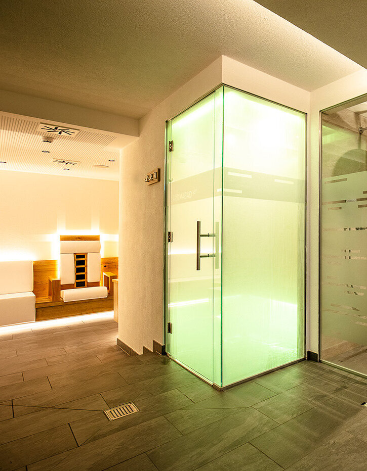 Modernes Spa-Interieur mit einem verglasten Dampfbad, das in grünem Licht erstrahlt, angrenzend an einen großen begehbaren Duschbereich mit Regenduschköpfen, beigen Fliesen und Milchglaselementen. Im Hintergrund ist eine Entspannungslounge zu sehen.