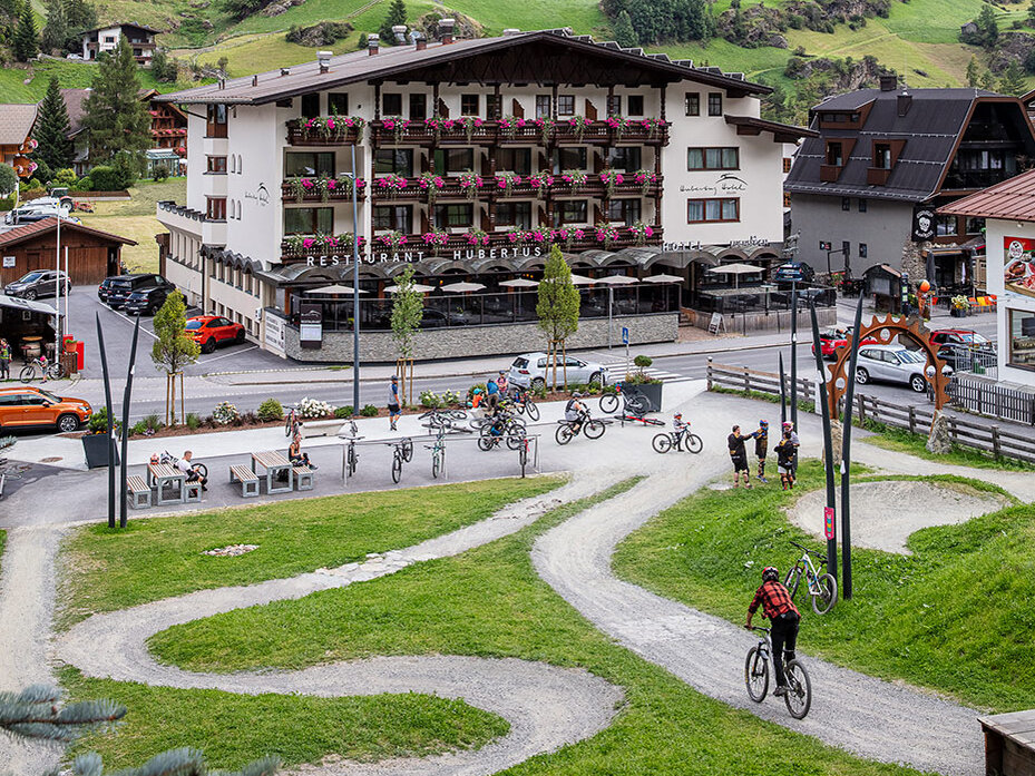 Ein malerisches Bergdorf mit einem großen Hotel, das über Balkone und Blumenkästen verfügt und von grünen Hügeln, Kiefern, gewundenen Radwegen und Radfahrern umgeben ist, die an einem sonnigen Tag unterwegs sind.