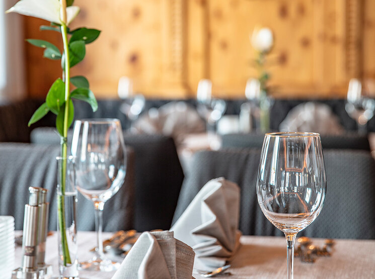 Elegant gedeckter Restauranttisch mit gefalteten grauen Servietten, Weingläsern, Besteck und einer einzelnen Blume mit grünem Stiel in einer Vase, vor einem unscharfen Hintergrund in warmen Farbtönen.