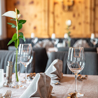 Elegant gedeckter Restauranttisch mit gefalteten grauen Servietten, Weingläsern, Besteck und einer einzelnen Blume mit grünem Stiel in einer Vase, vor einem unscharfen Hintergrund in warmen Farbtönen.