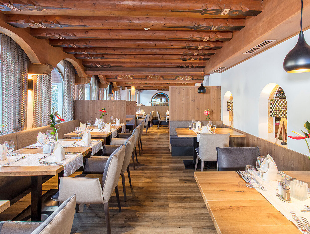 Ein gemütliches Restaurant mit Holzbalken, gedeckten Tischen mit weißen Servietten, moderner Beleuchtung und großen Bogenfenstern, durch die Tageslicht hereinströmt.