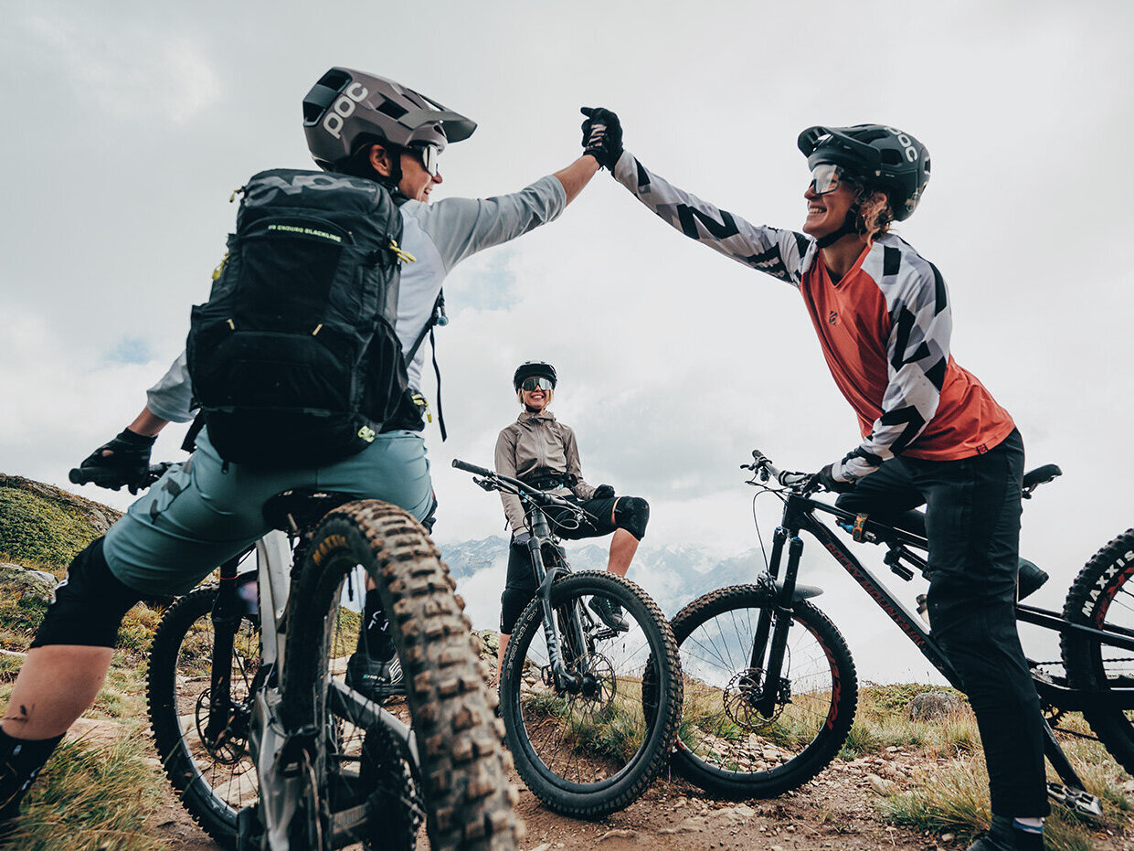 Drei Mountainbiker machen auf einem Trail eine Pause; zwei geben sich ein High-Five, während der dritte zusieht. Alle tragen Helme und Schutzausrüstung und sind von grasbewachsenen Hügeln unter einem bewölkten Himmel umgeben.