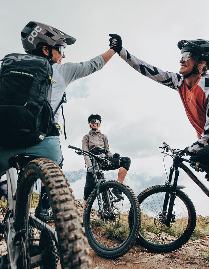 Drei Mountainbiker machen auf einem Trail eine Pause; zwei geben sich ein High-Five, während der dritte zusieht. Alle tragen Helme und Schutzausrüstung und sind von grasbewachsenen Hügeln unter einem bewölkten Himmel umgeben.
