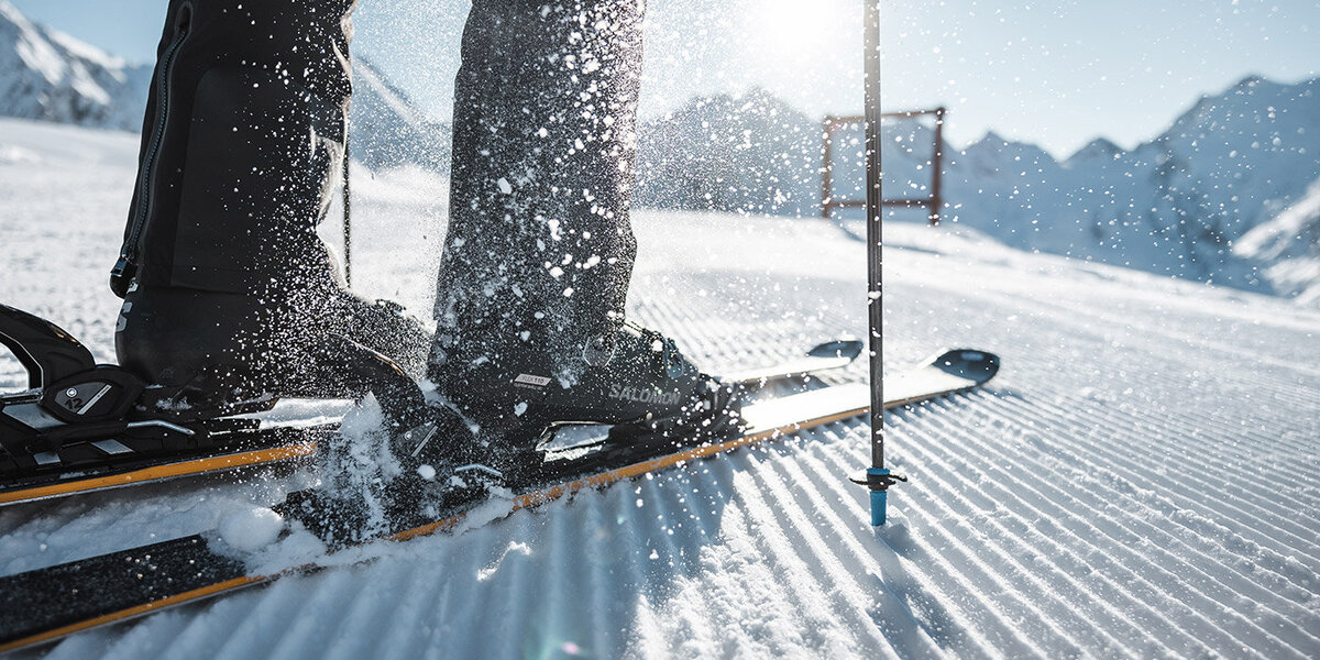 Nahaufnahme der Beine und Skier eines Skifahrers auf frisch präpariertem Schnee, mit Skistöcken und in die Luft spritzendem Schnee, vor einer strahlenden, sonnigen Bergkulisse.