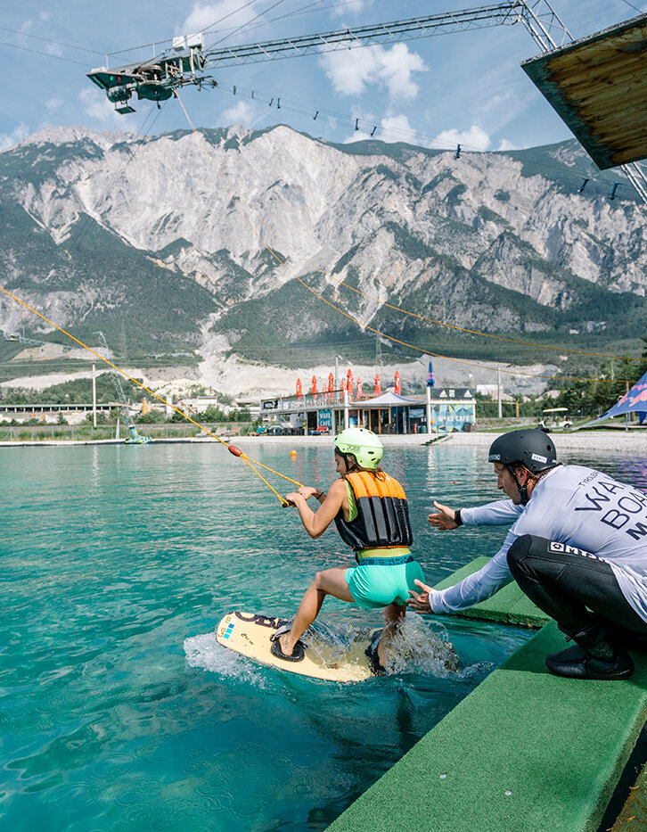 Eine Person auf einem Wakeboard macht sich bereit, von einem Steg aus zu starten, und hält dabei ein Seil fest. Eine weitere Person in Sportkleidung hilft ihr dabei. Im Hintergrund sind unter einem teilweise bewölkten Himmel Berge, Bäume und Gebäude zu sehen.