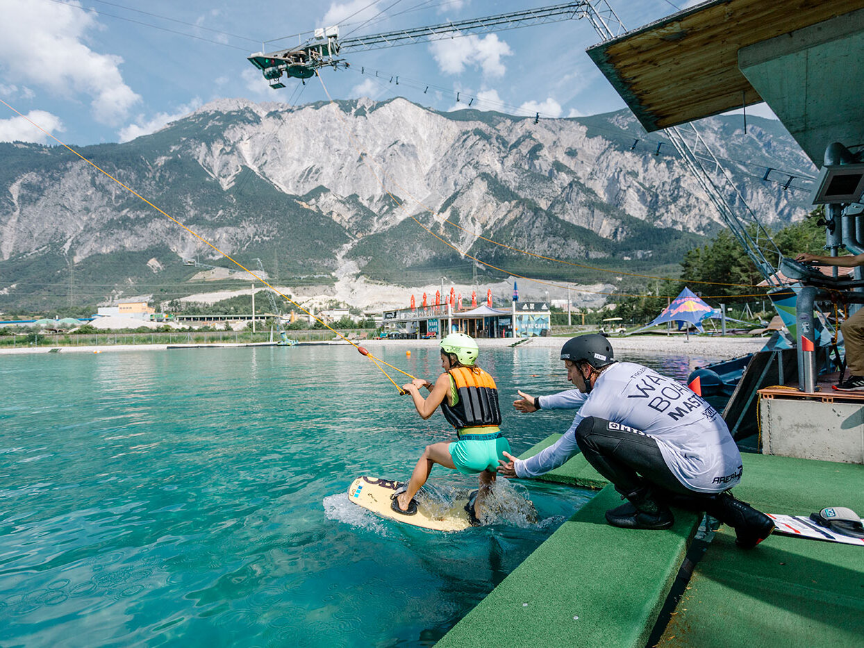Eine Person auf einem Wakeboard macht sich bereit, von einem Steg aus zu starten, und hält dabei ein Seil fest. Eine weitere Person in Sportkleidung hilft ihr dabei. Im Hintergrund sind unter einem teilweise bewölkten Himmel Berge, Bäume und Gebäude zu sehen.