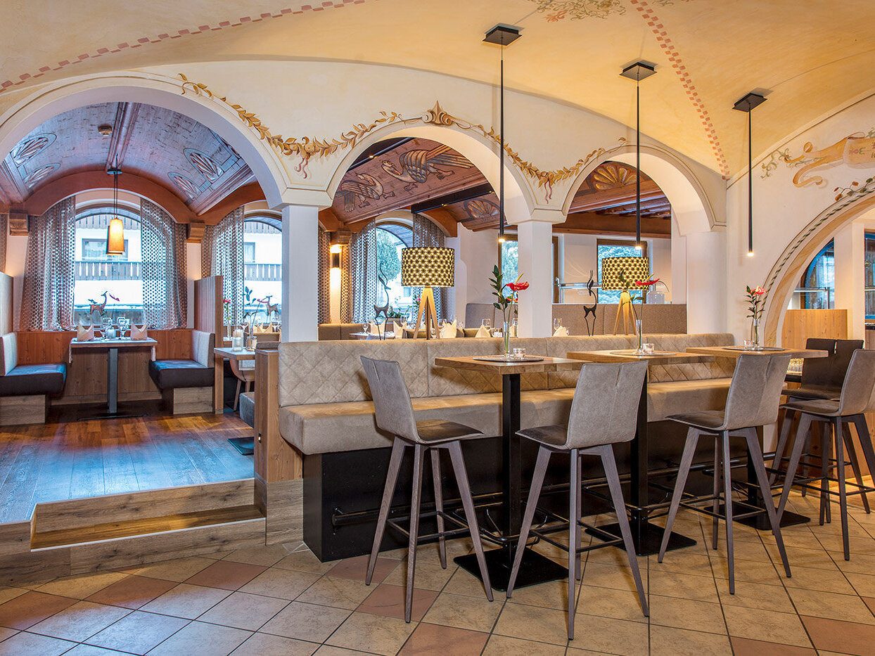 Elegantes Restaurantinterieur mit gewölbten Decken, gemütlichen Sitznischen am Fenster, einem zentralen Barbereich mit hohen Barhockern, warmer Beleuchtung und dekorativen Wandbildern. Die Atmosphäre ist einladend und stilvoll.