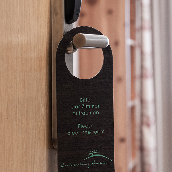 An der Türklinke einer Hotelzimmertür hängt ein Türhänger mit den Aufschriften „Bitte das Zimmer aufräumen“ und „Please clean the room“. Im Hintergrund sind Vorhänge und ein Teil des Zimmers zu sehen.