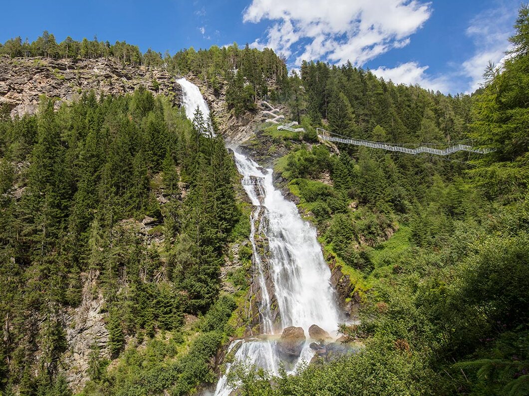 Ein hoher Wasserfall (Stuibenfall) stürzt einen bewaldeten Berg hinab, umgeben von üppig grünen Bäumen unter einem blauen Himmel mit vereinzelten Wolken. Eine Hängebrücke überspannt den oberen Teil des Wasserfalls.