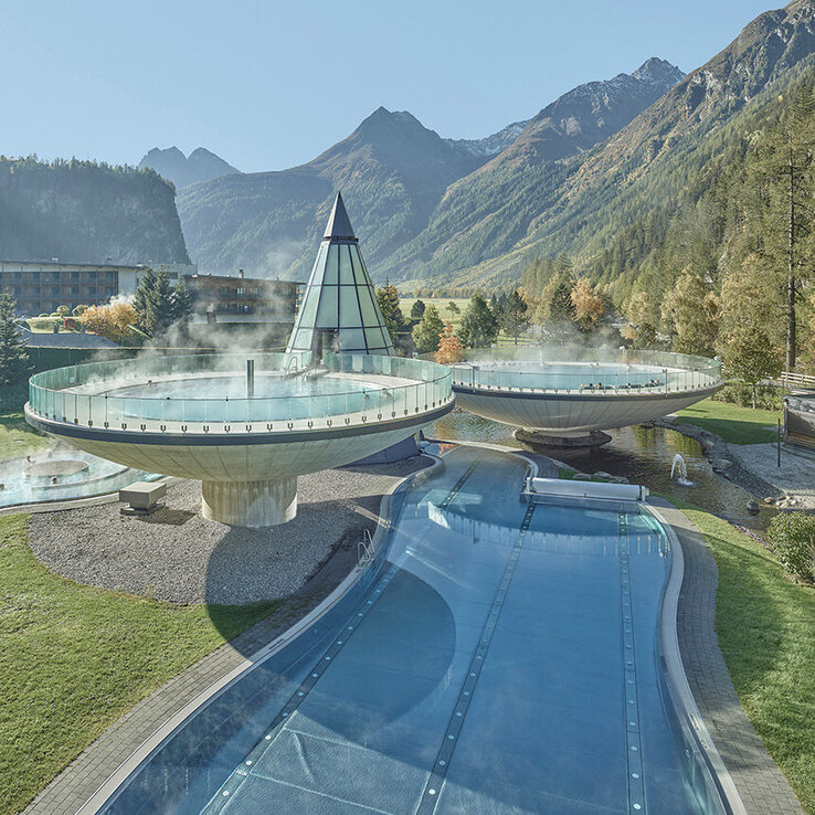 Ein modernes Thermalbad, vom Aqua Dome in Längenfeld, im Freien mit einer gläsernen Pyramidenkonstruktion, Dampfbecken, geschwungenen Wegen und umliegenden grünen Rasenflächen, vor der Kulisse bewaldeter Berge unter einem strahlend blauen Himmel.