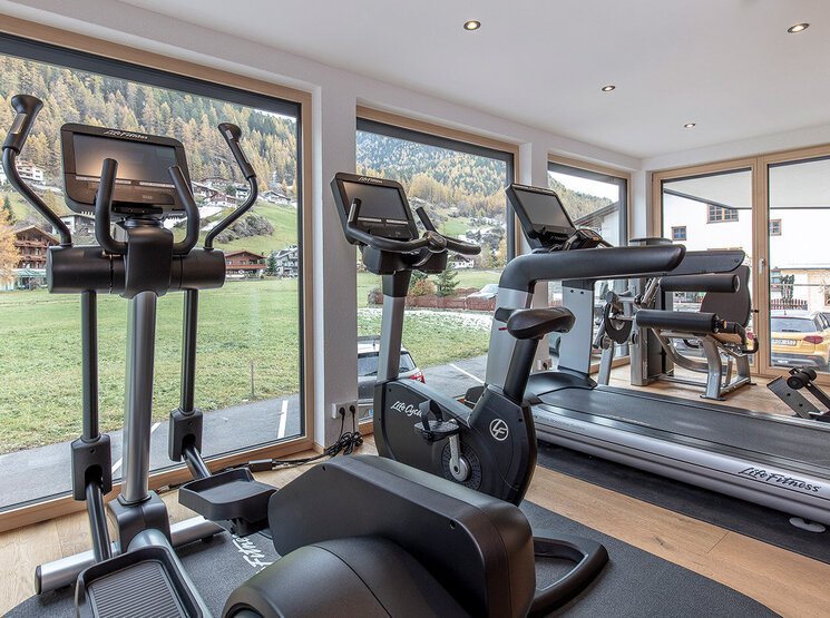 Ein modernes Fitnessstudio mit großen Fenstern, die einen malerischen Blick auf grüne Wiesen und Bäume bieten. Zur Ausstattung gehören stationäre Fahrräder, Crosstrainer und Laufbänder auf einem Holzboden. Der Raum ist von natürlichem Licht durchflutet.