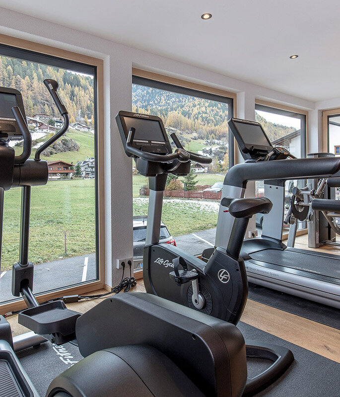Ein modernes Fitnessstudio mit großen Fenstern, die einen malerischen Blick auf grüne Wiesen und Bäume bieten. Zur Ausstattung gehören stationäre Fahrräder, Crosstrainer und Laufbänder auf einem Holzboden. Der Raum ist von natürlichem Licht durchflutet.