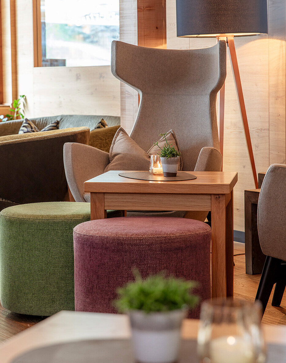 Eine gemütliche Lounge mit einem Sessel, runden grünen und violetten Sitzkissen, einem Holztisch, einer Stehlampe und Topfpflanzen auf den Tischen in einem sanft beleuchteten, modernen Raum.