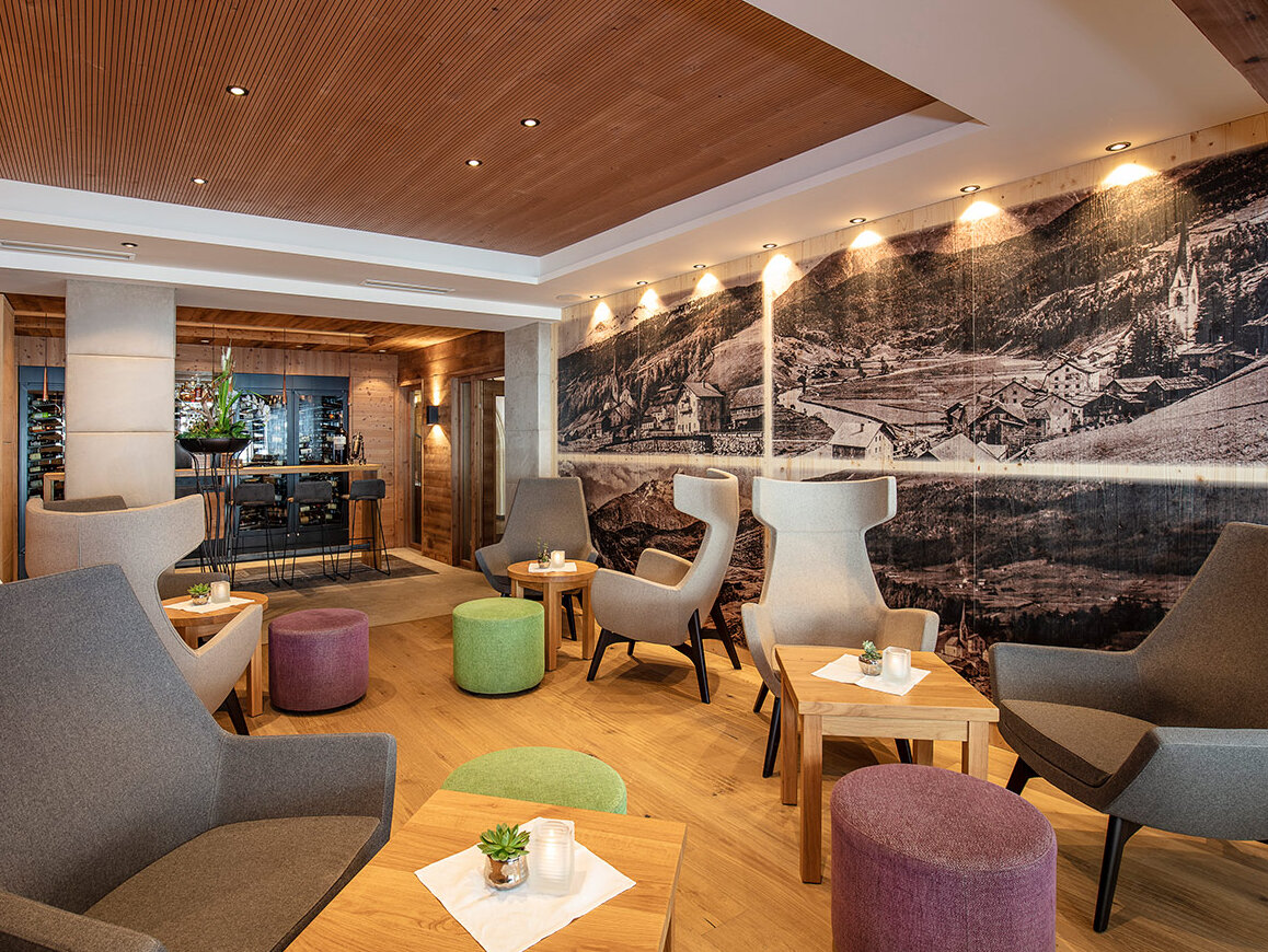 Moderne Lounge mit grauen Sesseln, kleinen runden Tischen und bunten Sitzkissen. Eine Wand ist mit einem schwarz-weißen Wandbild verziert. Der Raum ist warm beleuchtet, verfügt über Holzböden und -decken, und im Hintergrund ist ein Barbereich zu sehen.