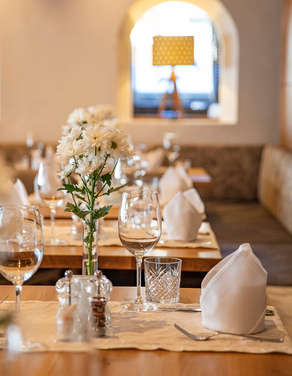 Ein gemütlicher Restauranttisch ist mit weißen Servietten, Gläsern, Besteck und einer Vase mit weißen Blumen gedeckt. Sanftes Licht und eine im Hintergrund verschwommen wirkende Lampe schaffen eine warme, einladende Atmosphäre.