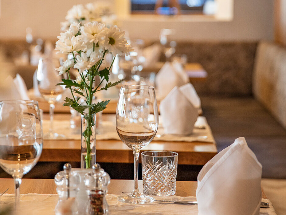 Ein gemütlicher Restauranttisch ist mit weißen Servietten, Gläsern, Besteck und einer Vase mit weißen Blumen gedeckt. Sanftes Licht und eine im Hintergrund verschwommen wirkende Lampe schaffen eine warme, einladende Atmosphäre.