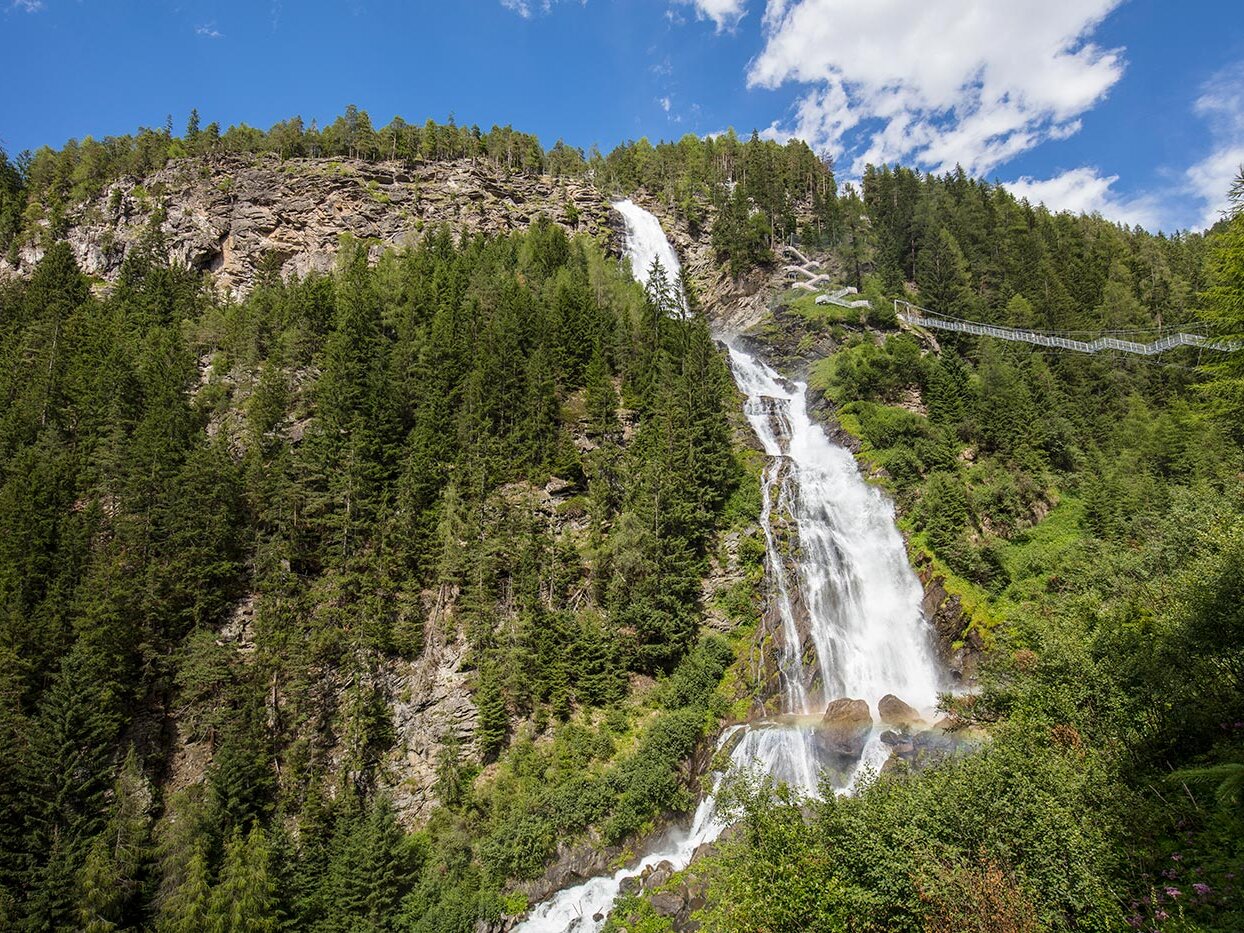 Ein hoher Wasserfall stürzt unter einem teilweise bewölkten blauen Himmel einen felsigen, bewaldeten Berghang hinab, umgeben von üppig grünen Bäumen und einer in der Ferne liegenden Fußgängerbrücke.