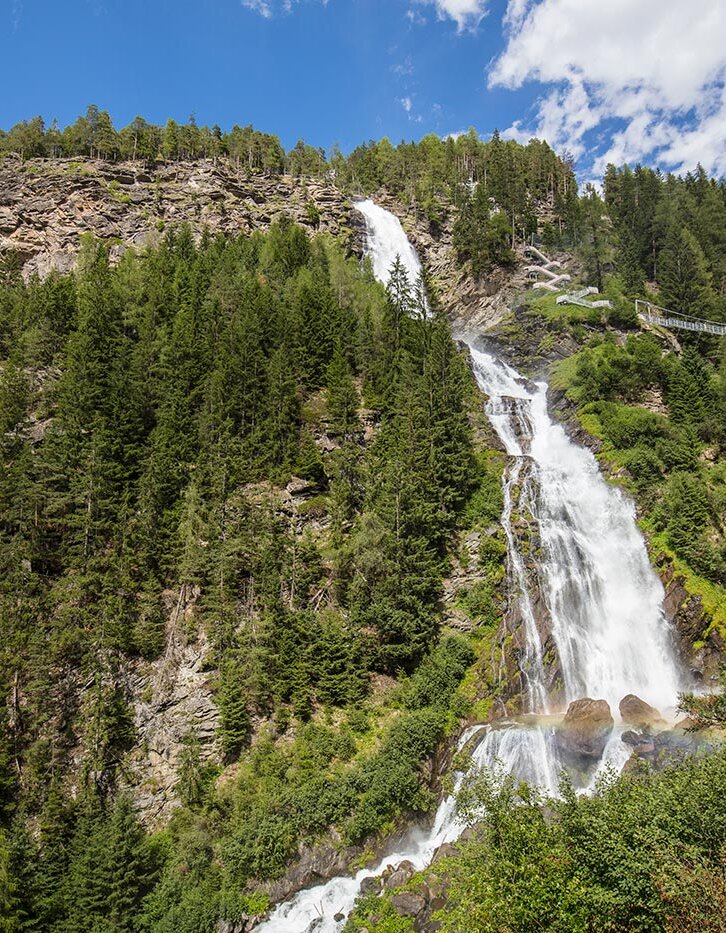 Ein hoher Wasserfall stürzt unter einem teilweise bewölkten blauen Himmel einen felsigen, bewaldeten Berghang hinab, umgeben von üppig grünen Bäumen und einer in der Ferne liegenden Fußgängerbrücke.
