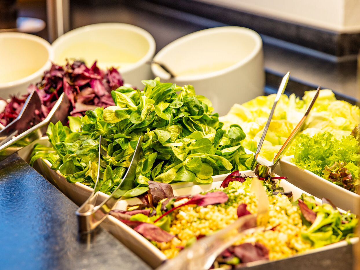 Eine Salatbar mit Behältern voller frischem Blattgemüse und gemischtem Salat sowie Zangen, im Hintergrund sind weiße Schüsseln zu sehen.