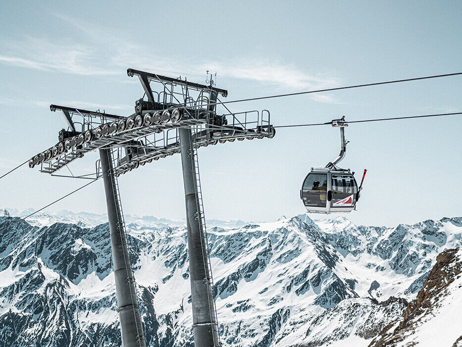 Eine Gondelbahn mit Fahrgästen fährt unter klarem Himmel über verschneite Berge, im Vordergrund ragen hohe Liftmasten empor, im Hintergrund sind schroffe Gipfel zu sehen.