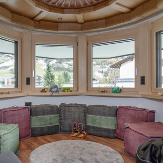 Gemütliches Spielzimmer mit großen Eckfenstern, die einen Blick auf die Berge bieten. Weiche Würfelsitzgelegenheiten umgeben einen runden Teppich, auf der Fensterbank liegen Holzspielzeug und Plüschtiere. Die warme Holzdecke und das natürliche Licht schaffen eine einladende Atmosphäre.