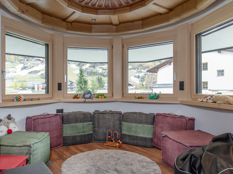 Gemütliches Spielzimmer mit großen Eckfenstern, die einen Blick auf die Berge bieten. Weiche Würfelsitzgelegenheiten umgeben einen runden Teppich, auf der Fensterbank liegen Holzspielzeug und Plüschtiere. Die warme Holzdecke und das natürliche Licht schaffen eine einladende Atmosphäre.