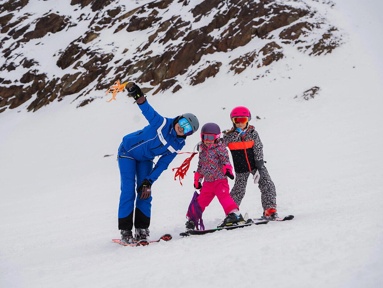 Ein Skilehrer in blauer Kleidung begleitet zwei Kinder in bunten Outfits auf einem verschneiten Hang; alle tragen Helme und Skibrillen, im Hintergrund sind felsige Berge zu sehen.