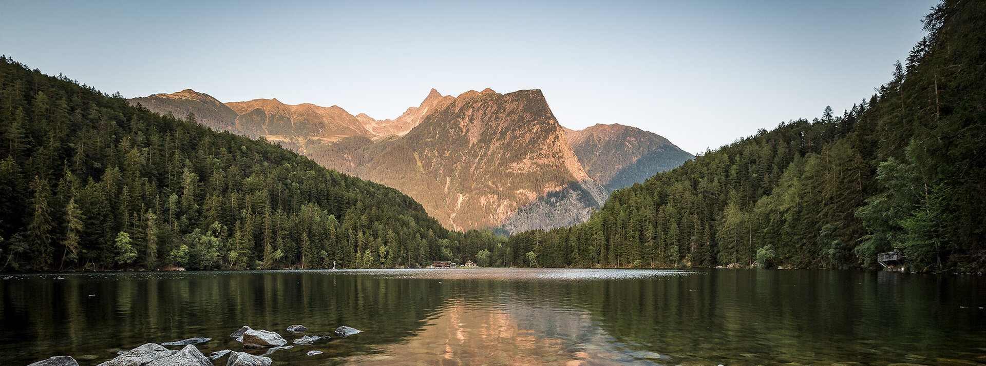 Das felsige Ufer des Piburger Sees, umgeben von dichtem, immergrünem Wald, mit fernen Bergen, die unter einem klaren Himmel in goldenes Sonnenlicht getaucht sind.