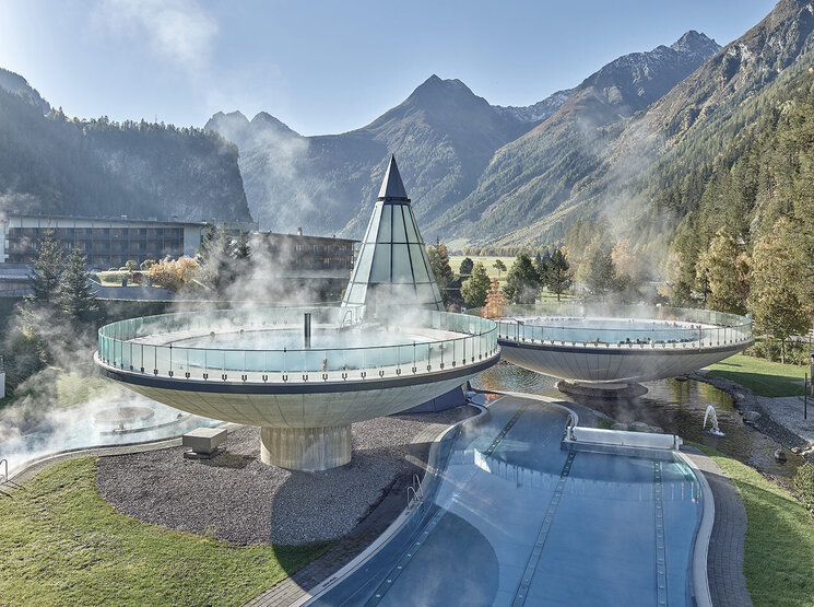 Dampfende, runde Außenpools mit Glasgeländern, eine gläserne Pyramidenkonstruktion, umgeben von Bergen und Bäumen an einem klaren Tag. Gezeigt wird der Aqua Dome in Längenfeld.