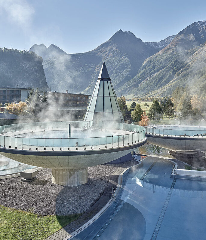 Dampfende, runde Außenpools mit Glasgeländern, eine gläserne Pyramidenkonstruktion, umgeben von Bergen und Bäumen an einem klaren Tag. Gezeigt wird der Aqua Dome in Längenfeld.