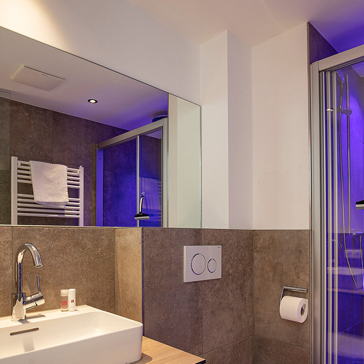 Modernes Badezimmer mit großem Spiegel, Waschbecken, Toilette und einer durch violette Beleuchtung beleuchteten Glasdusche. Handtücher und Pflegeprodukte sind ordentlich angeordnet, und die Einrichtung besticht durch Elemente aus Holz und Stein.