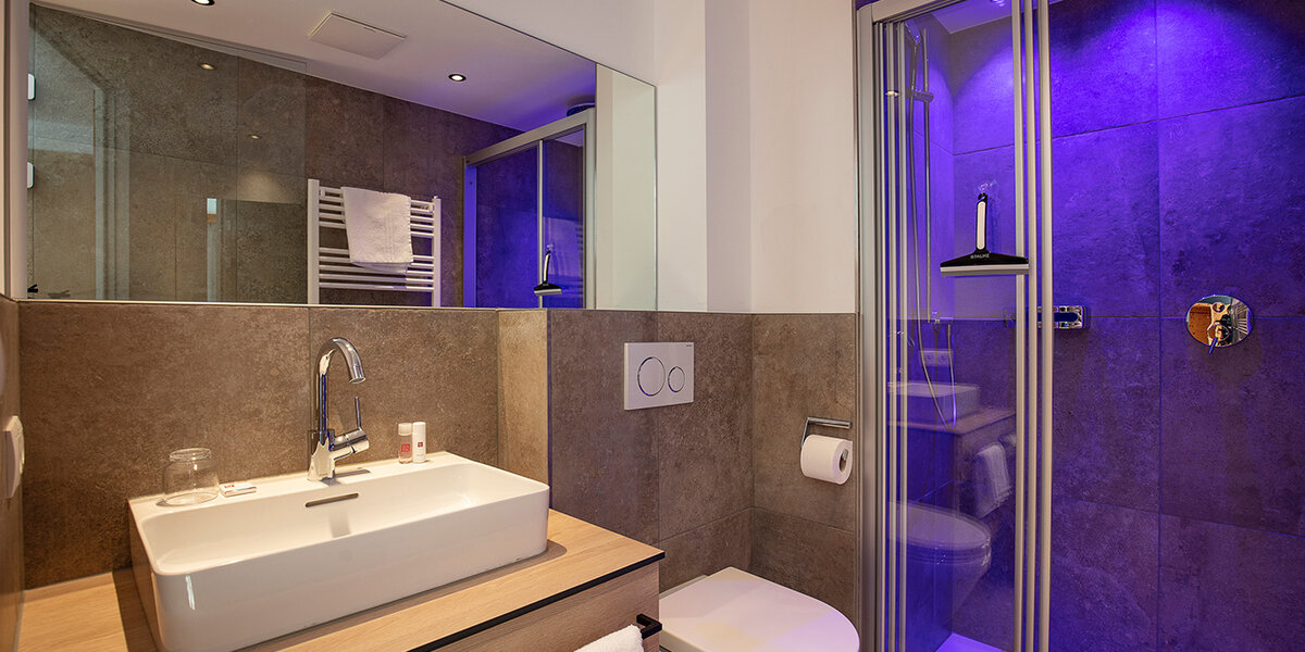 Modernes Badezimmer mit großem Spiegel, Waschbecken, Toilette und einer durch violette Beleuchtung beleuchteten Glasdusche. Handtücher und Pflegeprodukte sind ordentlich angeordnet, und die Einrichtung besticht durch Elemente aus Holz und Stein.