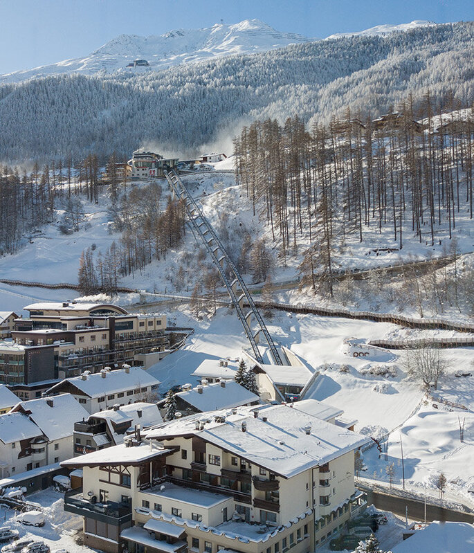 Ein verschneites Bergdorf mit Häusern, geparkten Autos und schneebedeckten Bäumen. Der Zentrum Shuttle führt hinter dem Hotel Hubertus den Hang hinauf, im Hintergrund sind Berge und ein strahlend blauer Himmel zu sehen.