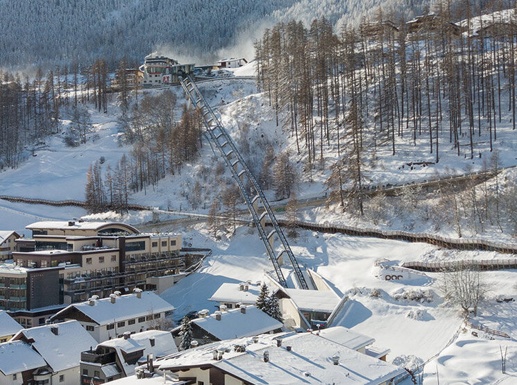 Ein verschneites Bergdorf mit Häusern, geparkten Autos und schneebedeckten Bäumen. Der Zentrum Shuttle führt hinter dem Hotel Hubertus den Hang hinauf, im Hintergrund sind Berge und ein strahlend blauer Himmel zu sehen.